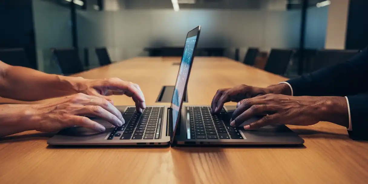 Two people reaching from opposite sides of an oak conference table toward a shared open laptop displaying a glowing dashboard, modern glass-walled office.