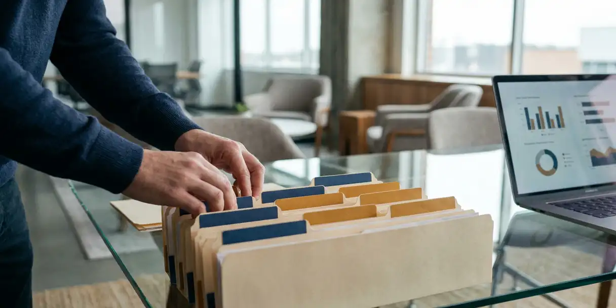 Data analyst organizing color-coded file folders on a glass desk beside a laptop displaying dashboard graphs in a modern office.